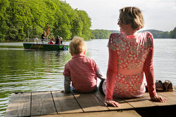 Mutter und Sohn sitzen auf einem Steg am Schmalen Luzin, Luzinfähre im Hintergrund, Naturpark Feldberger Seenlandschaft, Mecklenburg-Vorpommern, Deutschland