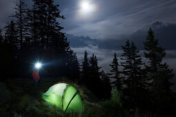Junge Frau mit Stirnlampe steht neben einem Zelt in einer Vollmondnacht, Bergell, Kanton Graubünden, Schweiz