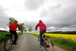 Two cyclists with electric bicycles passing blooming canola field, Tanna, Thuringia, Germany