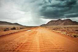 sandige rote Straße mit starkem Regen und Gewitter am Horizont, Tiras Gebirge, Tirasberge, Namib Naukluft Park, Namibia, Afrika