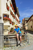 Radfahrerin rastet an einem Brunnen, Guarda, Unterengadin, Engadin, Kanton Graubünden, Schweiz
