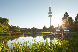 People sitting on the terrace of the Seepavillon snack bar at the park lake, television tower in the background, Planten un Blomen, Hamburg, Germany