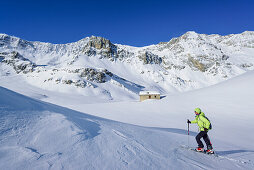 Frau auf Skitour steigt an verschneiter Almhütte vorbei mit Monte Reghetta, Monte Faraut und Monte Gabel im Hintergrund, Rocca La Marchisa, Valle Varaita, Cottische Alpen, Piemont, Italien