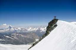 Two female alpinists holding on to the summit cross of Allalinhorn and waving, in the background from left to right Matterhorn, Dent Blanche and Zinalrothorn, Pennine Alps, canton of Valais, Switzerland