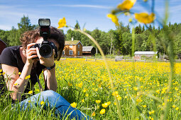 Junge Frau sitzt in einer Wiese mit Butterblumen und fotografiert mit einer Nikon Kamera, Trondheim, Sør-Trøndelag, Norwegen, Europa