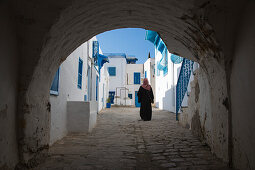 Arabian woman walks through archway along path of buildings with blue windows and white walls in artisan village, Sidi Bou Said, Tunis, Tunisia