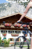 Young woman riding past a house on her bike on a sunny day, Tannheimer Tal, Tyrol, Austria