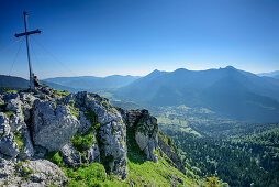 Person sitzt am Gipfelkreuz des Leonhardstein, Blick auf Tegernseer Berge, Leonhardstein, Bayerische Alpen, Oberbayern, Bayern, Deutschland