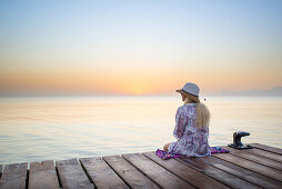 Young blond-haired woman sitting on the end of a long pier in the morning mood, enjoying the view of the sunrise. Playa de Muro beach, Alcudia, Mallorca, Balearic Islands, Spain