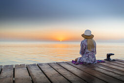 Young blond-haired woman sitting on the end of a long pier in the morning mood, enjoying the view of the sunrise. Playa de Muro beach, Alcudia, Mallorca, Balearic Islands, Spain