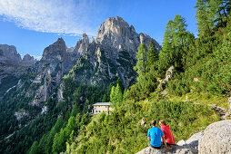 Two persons hiking sitting on rock in front of hut Rifugio Treviso with Cima dei Lastei, valley Val Canali, Pala range, Dolomites, UNESCO World Heritage Dolomites, Trentino, Italy