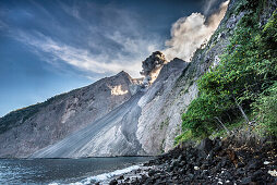 Großen Asche-Eruption und Gesteinsauswurf an der offenen Flanke mit Feuerrutsche des Vulkans Batu Tara mit austretenden Vulkan-Gasen und Asche in der Flores Sea bei Tage vor bleuem Himmel mit steiniger Küste, Meer, und Vegetation im Vordergrund, Insel Kom