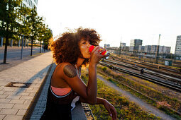 Young afro-american woman drinking from a can in urban scenery with train tracks, Hackerbruecke Munich, Bavaria, Germany