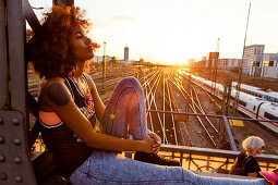 Junge afroamerikanische Frau sitzt im Abendlicht auf Stahlträger in urbaner Szenerie mit Gleisen, Hackerbrücke, München, Bayern, Deutschland