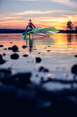 Junger Windsurfer traegt sein Segel und Surfbrett aus dem Wasser bei Sonnenuntergang, Ammersee, Bayern, Deutschland