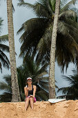 Junge Surferin sitzt im Sand und isst eine Kokosnuss, Sao Tome, Sao Tome und Príncipe, Afrika
