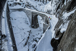 Zwei Junge Skifahrer gehen über eine verschneite Steinbrücke, Andermatt, Uri, Schweiz