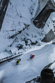 Zwei Junge Skifahrer fahren auf einer verschneiten Straße bei einer Steinbrücke, Andermatt, Uri, Schweiz