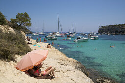 People on rocks relax under sun umbrella with yachts and sailboats at anchor in Cala Portals Vells bay, Portals Vells, Mallorca, Balearic Islands, Spain