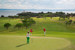 Golfers on green of hole 12 at Club de Golf Alcanada with Faro de Alcanada lighthouse in distance, near Port d'Alcudia, Mallorca, Balearic Islands, Spain