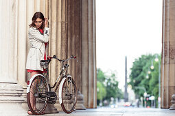 Young woman with her bicycle at dusk underneath the propylae, Königs Plaza, overlooking the Obelisk, Munich, Bavaria, Germany