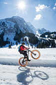 Ein junger Mann zeigt ein Wheelie auf einem Fatbike, Snowbike, Mountainbike vor dem zugefrorenen Lauenensee (Louwenesee) in der Nähe von Gstaad, Berner Oberland, Schweiz