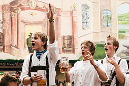 Young men in leather trousers standing on beer benches celebrate Oktoberfest in the beer tent