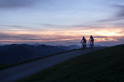 Zwei junge Männer auf ihren Rennrädern bei Sonnenuntergang in den Kitzbühler Alpen, Kitzbühlerhorn, Tirol, Österreich