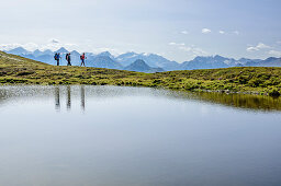 Three persons hiking alongside lake, High Tauern in background, Pinzgau walk, Kitzbuehel Alps, Salzburg, Austria