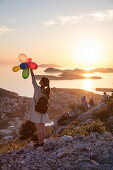 Young Asian woman holds colorful balloons and admires view across Lapad Peninsula and islands seen from hillside near top of Dubrovnik Gondola at sunset, Dubrovnik, Dubrovnik-Neretva, Croatia