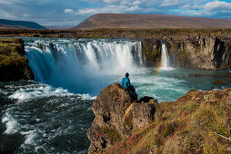 A tourist sits on a rock promontory, viewing the 12 meter high Goðafoss (Waterfall of the Gods), near Seyðisfjörðdur, Eastern Iceland, Europe