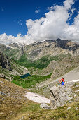 Woman hiking sitting on rock and looking towards valley, mountain lake and Cottian Alps in background, Val Maira, Cottian Alps, Piedmont, Italy