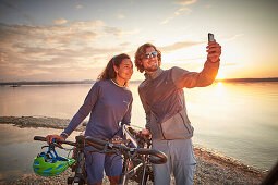 junge Frau auf Tourenrad und junger Mann auf eTourenfahrrad, machen ein selfie am Kiesstrand, Radtour am See, Münsing, Bayern, Deutschland
