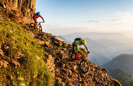 Two middle aged men ride down a rocky and steep hiking path on their mountainbikes, morning light, Wilder Kaiser mountain range in the background, Kirchberg, Tyrol, Austria