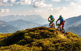 Zwei Mountainbiker fahren über einen bewachsenen Hügel in den Kitzbühler Alpen, Wilder Kaiser im Hintergrund, Kirchberg, Tirol, Österreich