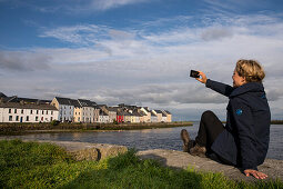Eine blonde Frau in einer blauer Jacke und Wanderschuhen macht ein Selfie Foto mit dem Straßenzug The Long Walk – einer Reihe von farbenfrohen Häusern im Hafen von Corrib – im Hintergund, Galway, County Galway, Irland, Europa