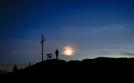 Person at the summit cross of the Jochberg at night with clouds, stars and moon