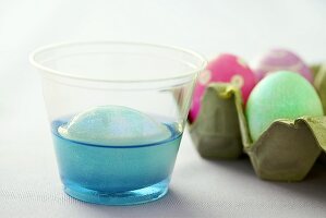Ei im Becher mit blauer Eierfarbe neben gefärbten Eiern