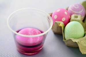 Ei im Becher mit lila Eierfarbe neben gefärbten Eiern