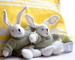 Two white Easter bunny with self-made knitted sweaters on yellow cushion