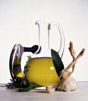 Glaskaraffe mit Olivenöl, daneben Knoblauch, Kräuter u. Wiegemesser