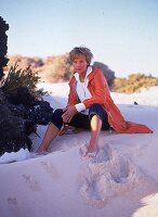 Frau sitzt im Sand, Capri-Jeans, orangeroter, knielanger Parka