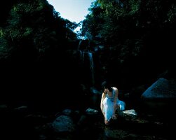 Frau in weißem langem Wickelrock und Top hockt vor einem Wasserfall