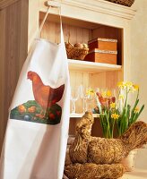 Easter apron with straw hen and daffodils in vase in front of cabinet