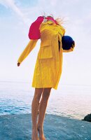 Frau im gelben Mantel am Meer springt in die Luft, hält Ball