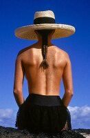 Rear view of semi dressed woman with long fish tail braid wearing sunhat sitting on rocks