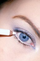 Close-up of blue eyed woman wearing eye shadow applying white eyeliner with pencil