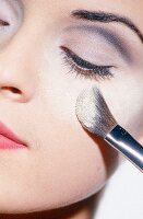 Close-up of woman wearing gray eye shadow applying powder on cheeks with brush