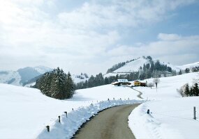 Schneebdeckter Berg, davor zwei Häuse, Weg schlängelt sich hinauf