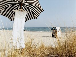 Rüschenkleid am Kleiderbügel unter Sonnenschirm am Strand, Strandkorb